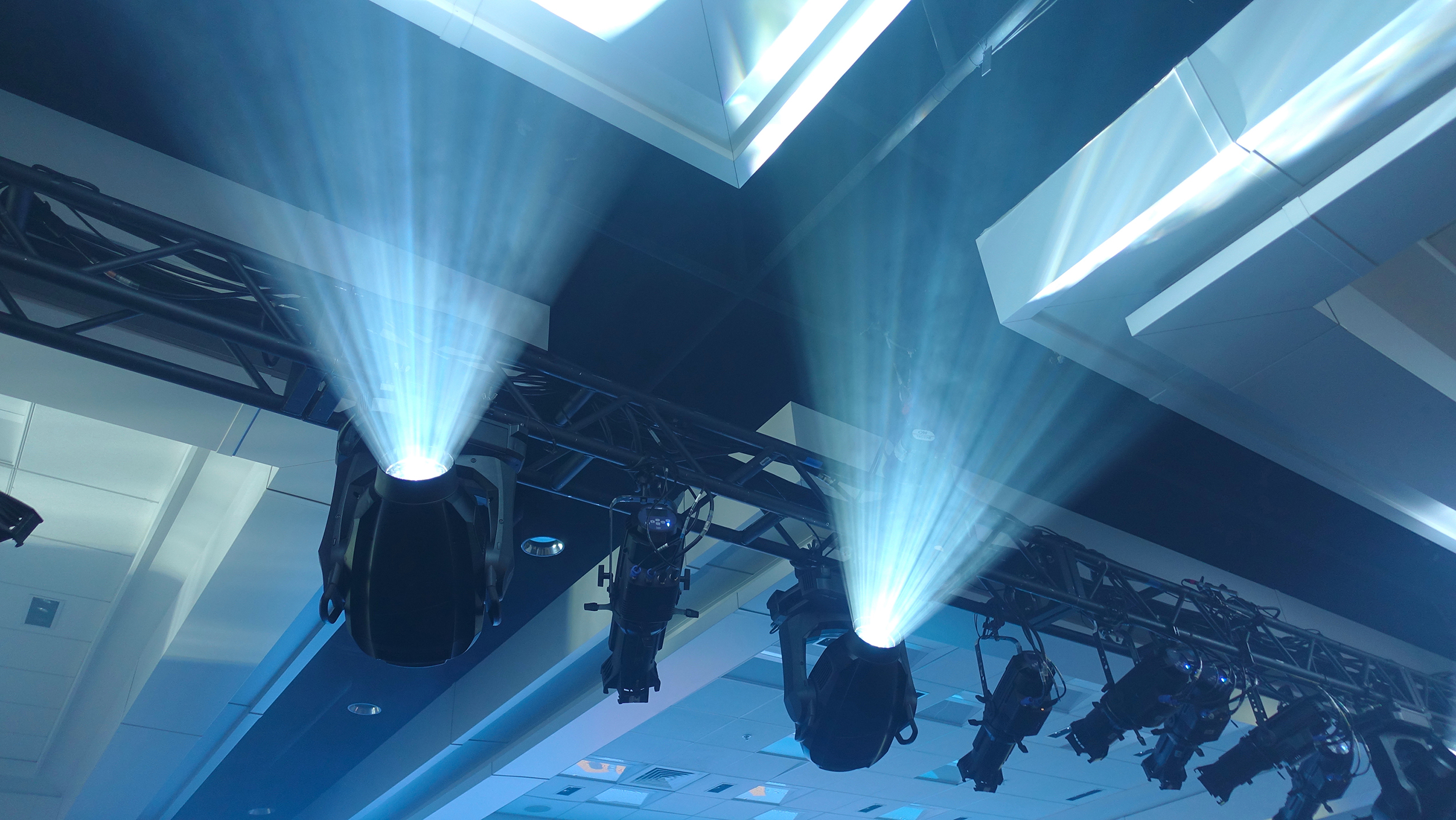 Stage lights mounted on a metal truss beam shine bright white beams upward in an indoor venue with a high ceiling and geometric panels, creating a dramatic lighting effect.