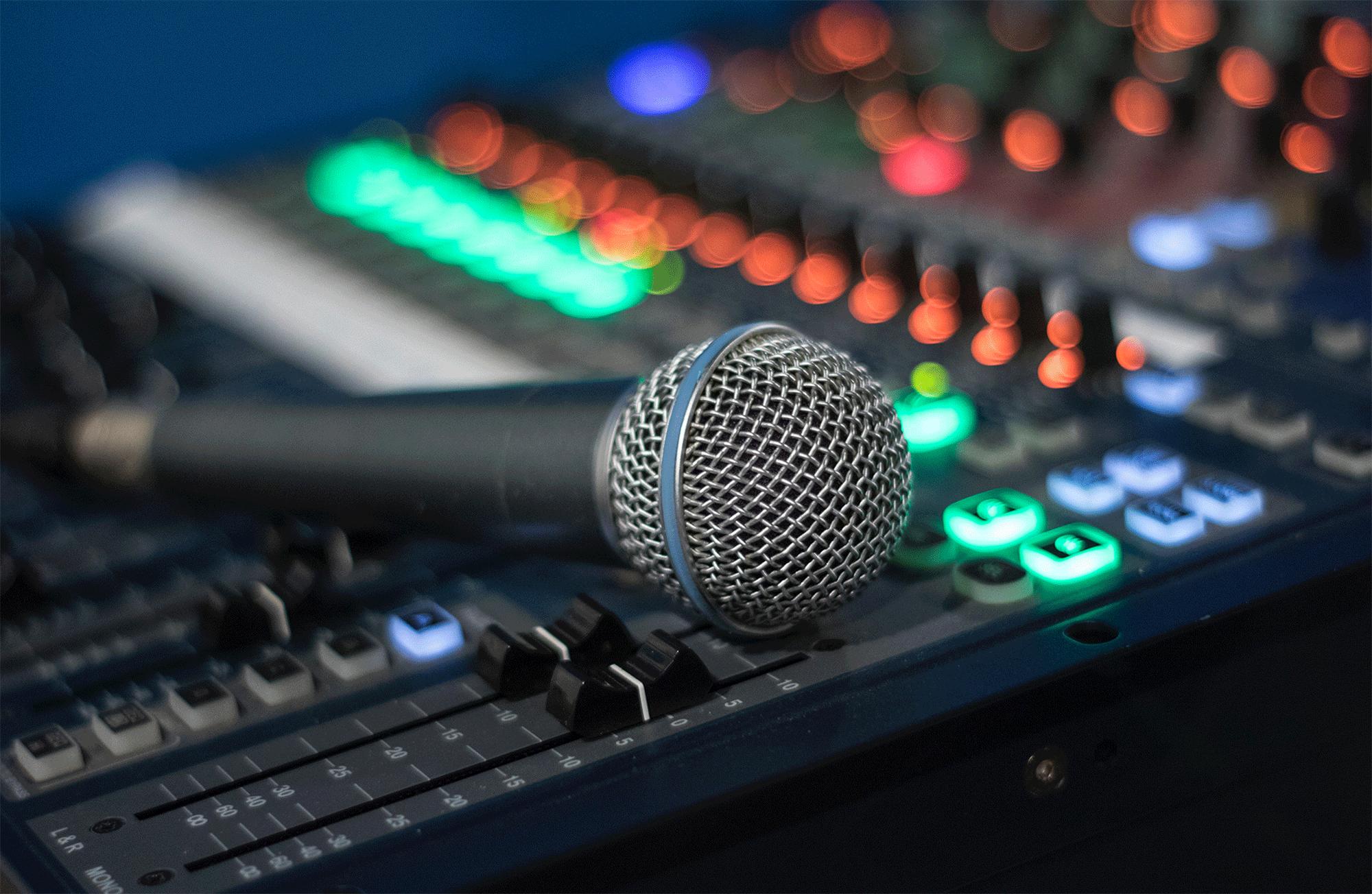 A close-up of a microphone resting on a sound mixing board with illuminated buttons and sliders, creating a colorful and vibrant background.