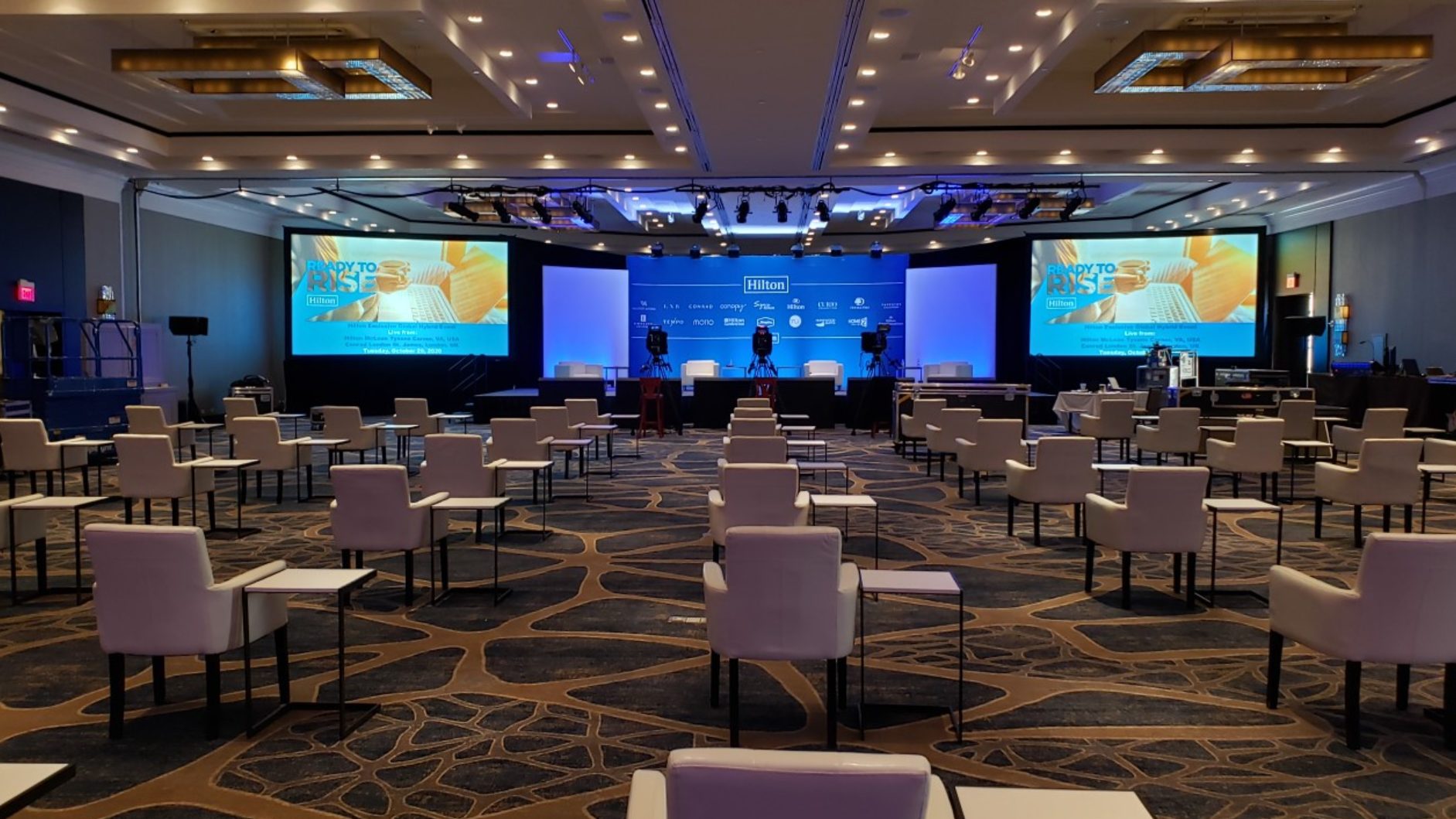 A large conference room with rows of white chairs spaced apart facing a stage with three chairs, cameras, bright lights, and two large screens displaying the words “Unstoppable Rise.”.