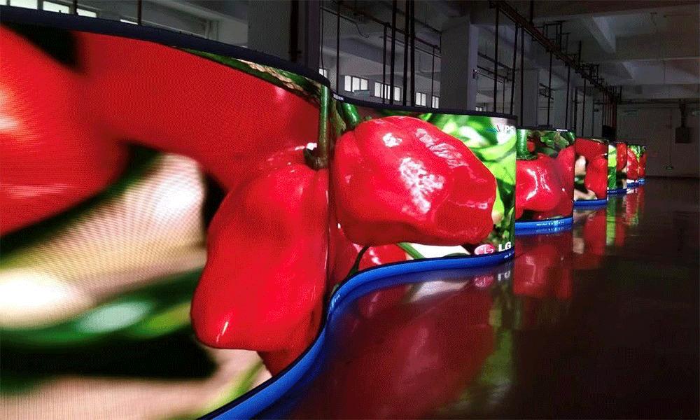Curved LED screens display vibrant close-up images of red peppers in a large indoor space with reflective floors and exposed ceiling pipes.
