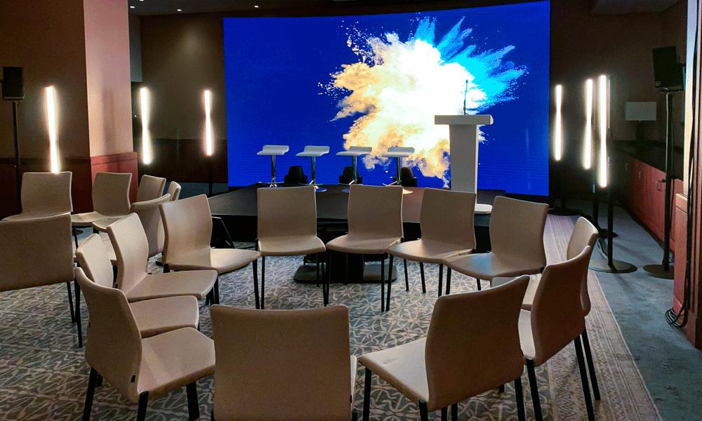 A conference room with white chairs arranged in a semicircle facing a stage, which has a podium, three stools, and a large digital screen displaying a colorful explosion graphic. Vertical lights stand on either side of the stage.