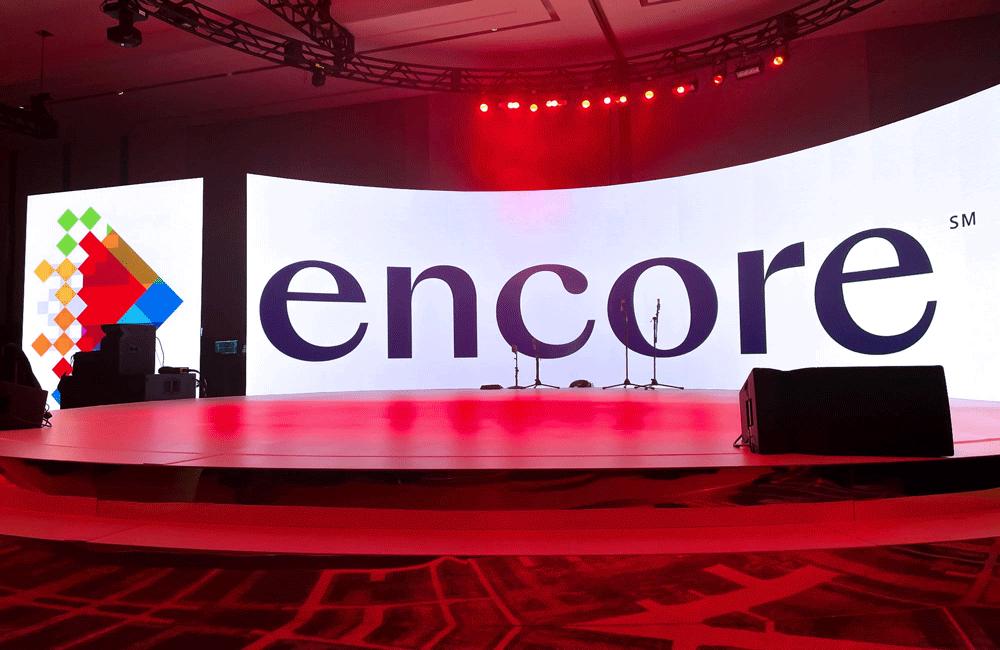 A brightly lit stage with a large curved LED screen displaying the word encore and a colorful geometric logo. Red lights shine from above, and microphones are set up at the center of the stage.