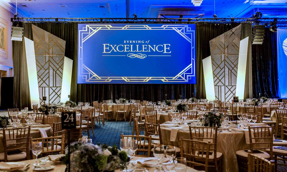 A large banquet hall decorated for an event, with round tables set for dinner and a big blue screen at the front that reads Evening of Excellence in elegant gold lettering.