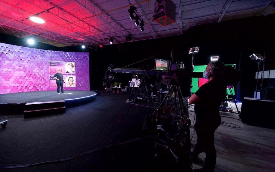 A person stands on a stage with a hexagonal-patterned backdrop, facing a large video camera and crew in a studio with bright lighting and screens displaying images and text.