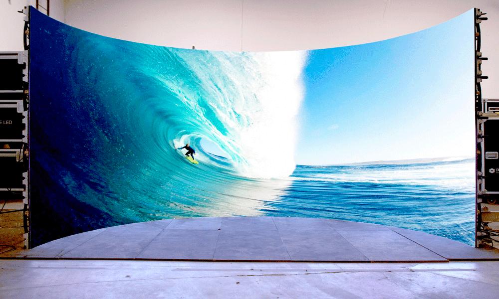 A large, curved LED screen displays a vivid image of a surfer riding inside a powerful blue wave, set up indoors on a bare, curved platform with visible equipment in the background.
