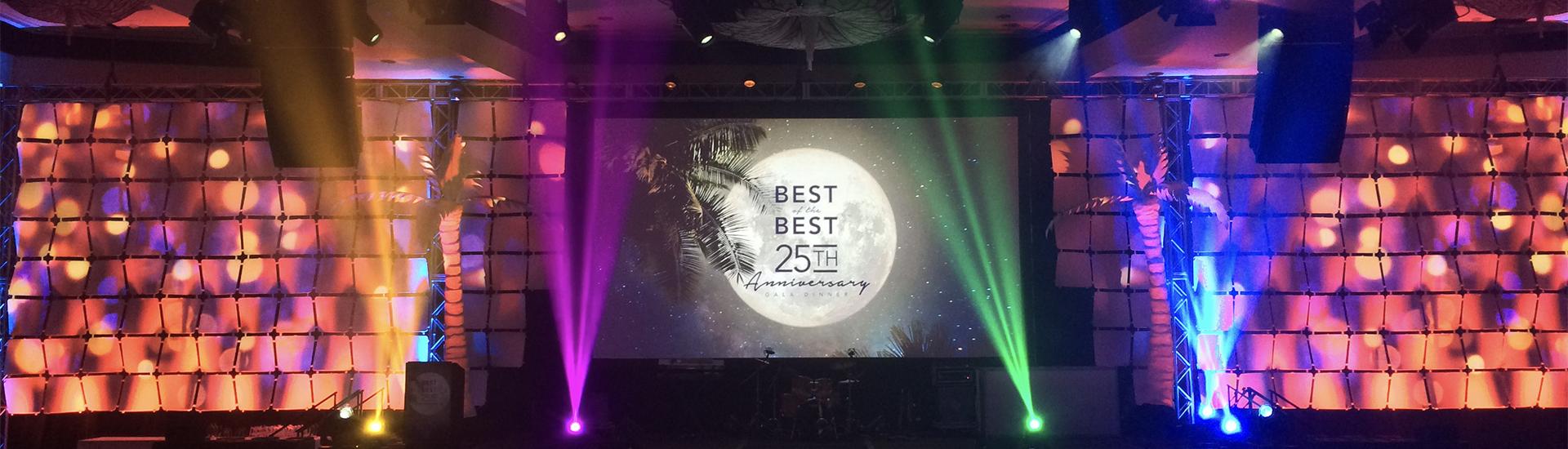 A stage decorated with colorful spotlights and palm tree silhouettes features a large screen reading Best of the Best 25th Anniversary against a bright moon backdrop.
