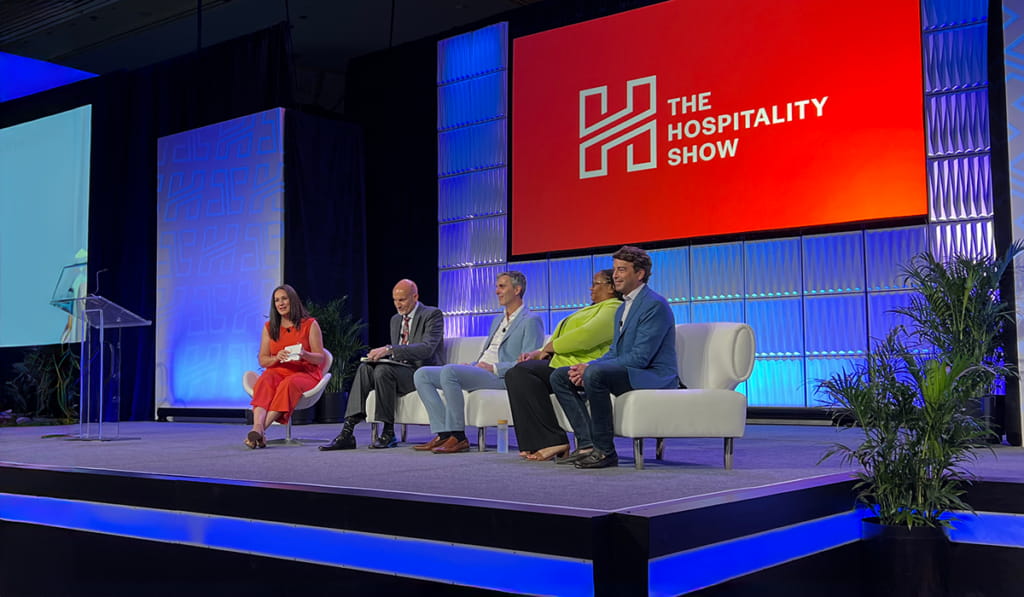 Five people sit on stage for a panel discussion at The Hospitality Show event, with a large red screen displaying the event logo behind them and decorative plants on either side of the stage.