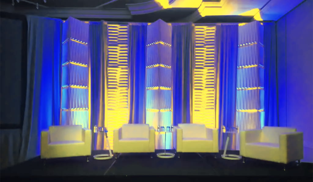 A stage setup with four white armchairs, small round side tables, and a backdrop illuminated with blue and yellow lighting patterns. The background features vertical textured panels and draped curtains.