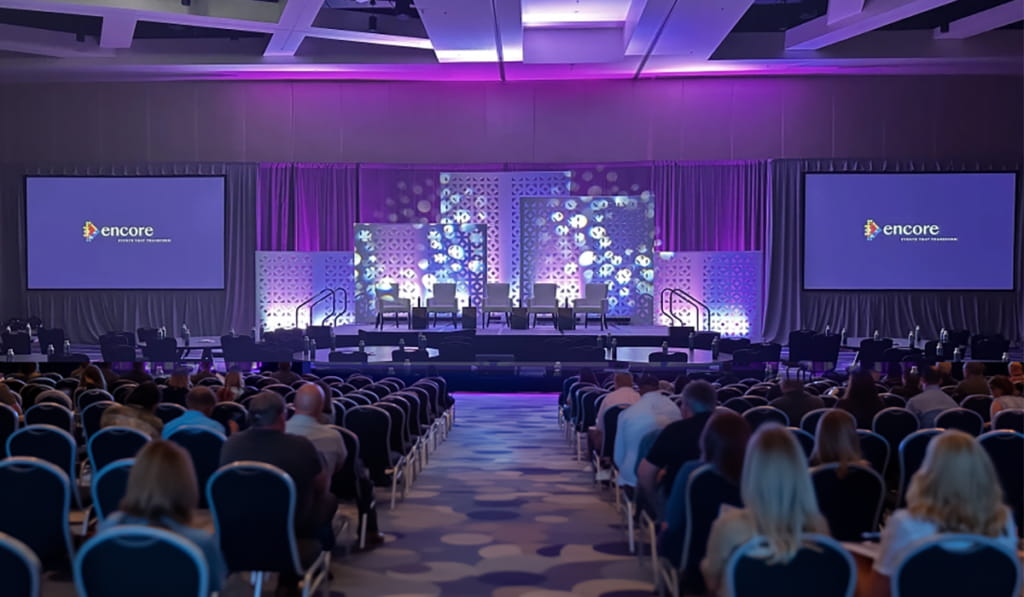 A large conference room filled with seated attendees faces a stage with four chairs, two large screens displaying the Encore logo, and purple ambient lighting.