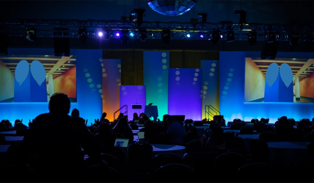 A dimly lit conference room with people seated at round tables, facing a colorful stage with blue, orange, and purple lighting, large screens, and decorative circular patterns.