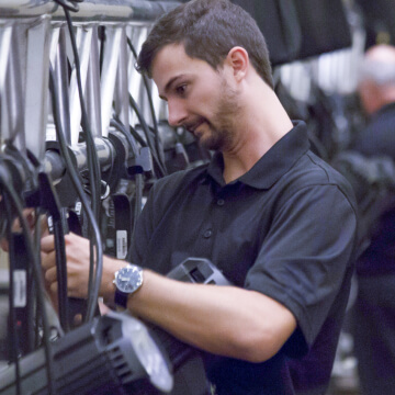 A man in a black polo shirt adjusts stage lighting equipment, focusing intently as he works with cables and fixtures in a professional setting.