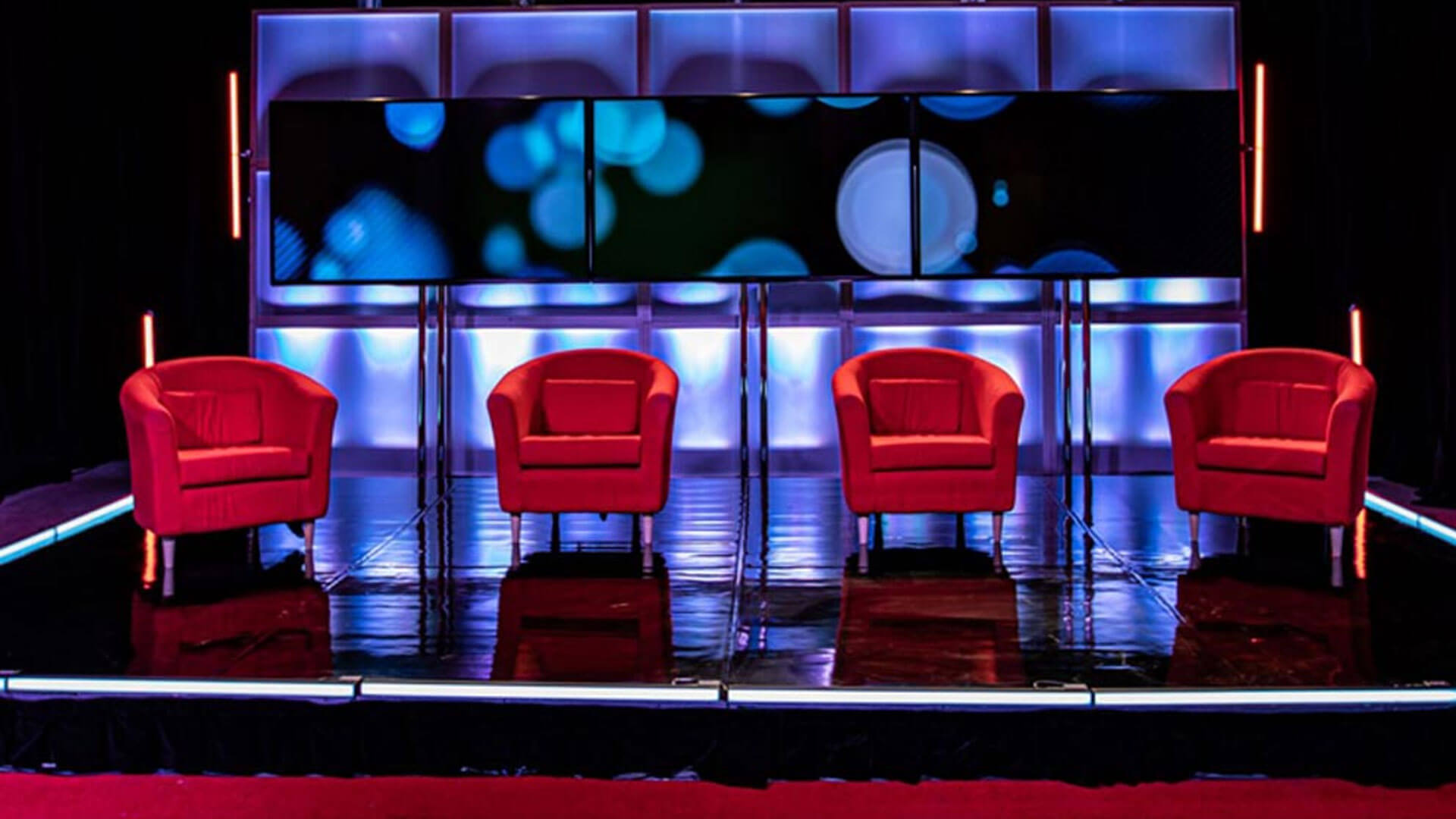 Four empty red armchairs are arranged in a row on a shiny black stage, in front of a large digital screen with abstract blue and white light patterns, with vertical lights on each side.