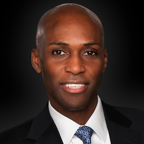 A man with a shaved head wearing a dark suit, white shirt, and blue patterned tie, smiling against a black background.