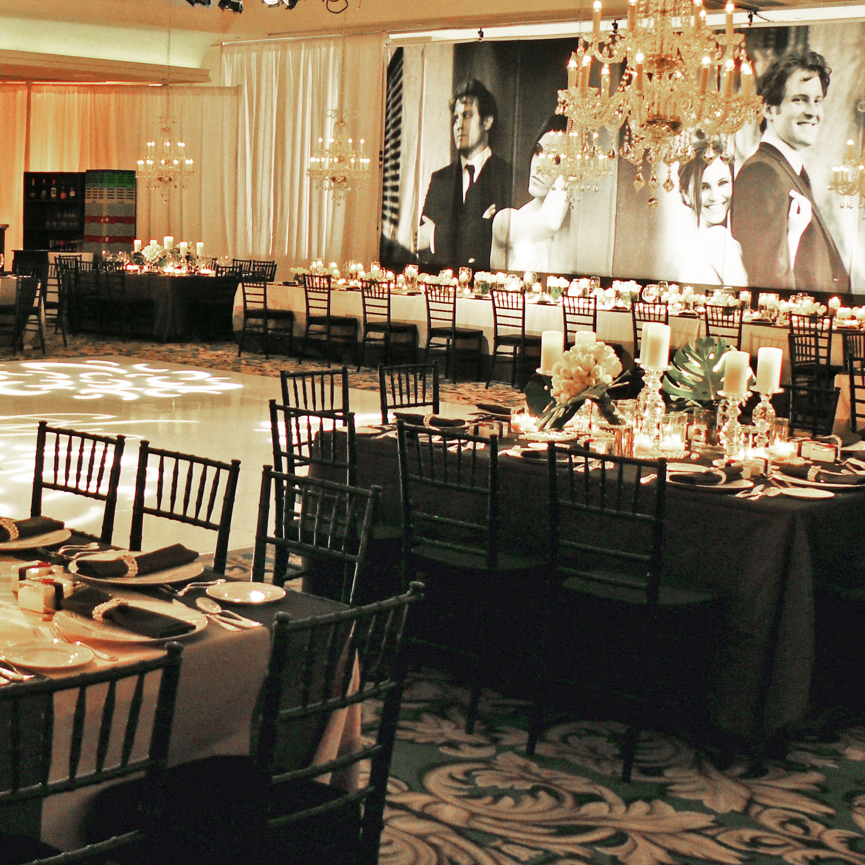An elegant banquet hall set for an event, with round and rectangular tables, black chairs, chandeliers, floral centerpieces, and a large black-and-white wall photo of a bride and groom.