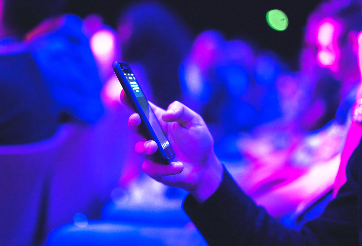A person holds a smartphone in one hand, surrounded by a blurred crowd, under vibrant blue and purple lighting, possibly at an event or conference.