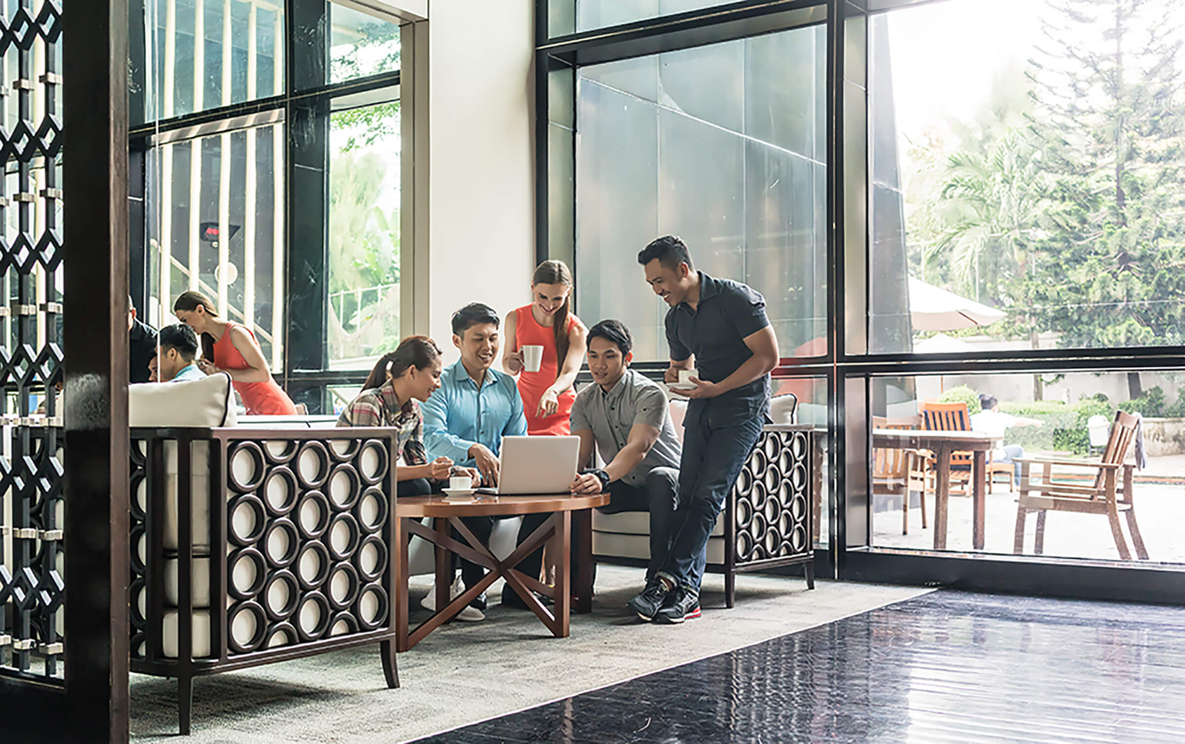 Five people gather around a table in a modern, sunlit lounge. Four people sit, engaged with a laptop, while a server brings drinks. Large windows reveal a green outdoor area with trees and patio furniture.